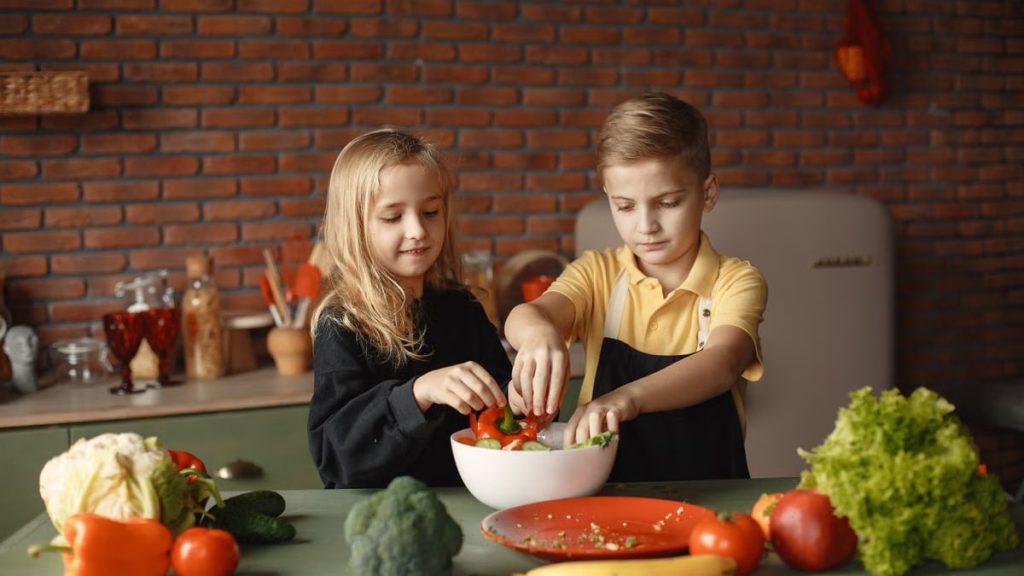 Cheerful girl helping little boy in apron preparing salad while standing in kitchen near table with mix of fresh vegetables behind brick wall